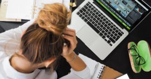 Overhead view of a stressed woman working at a desk with a laptop, phone, and notebooks overwhelmed with executor responsibilities.